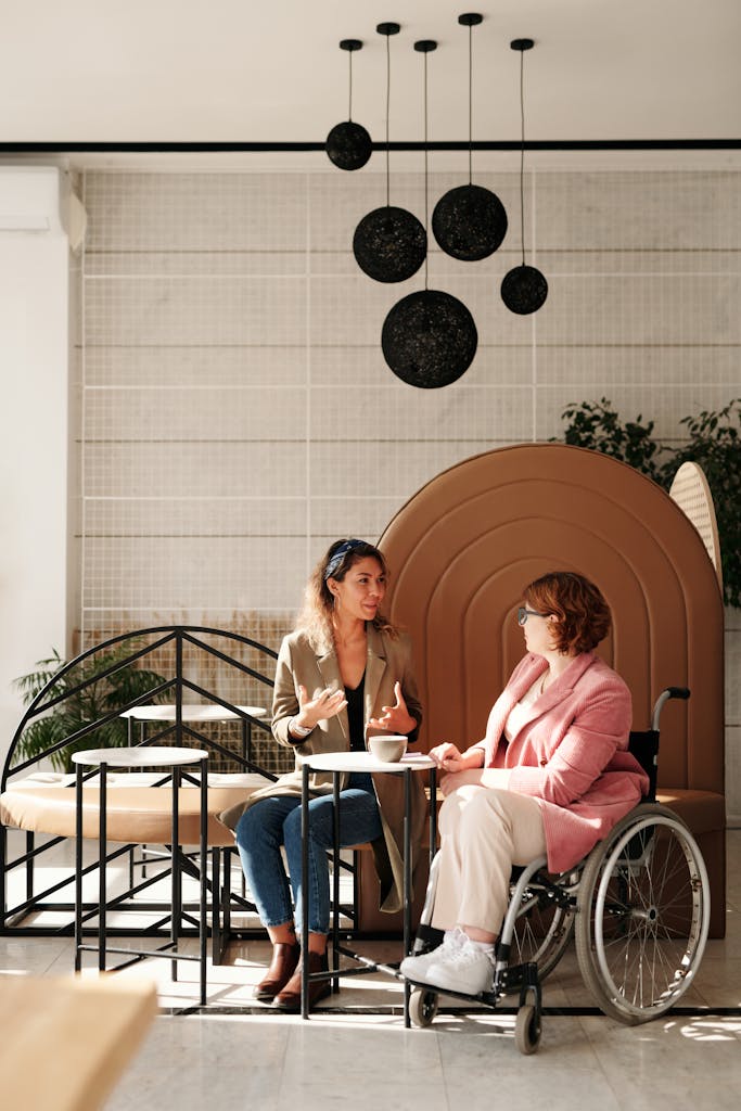 Two women share a conversation, one seated in a wheelchair, in a stylish cafe.